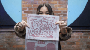 Brick background with half light blue and half dark blue frames on each side. In the center the artist is holding uo their artwork.